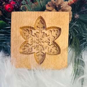 May include: A wooden cookie mold with a carved snowflake design. The mold is square and made of light-colored wood. The snowflake is detailed with intricate patterns, perfect for holiday baking.