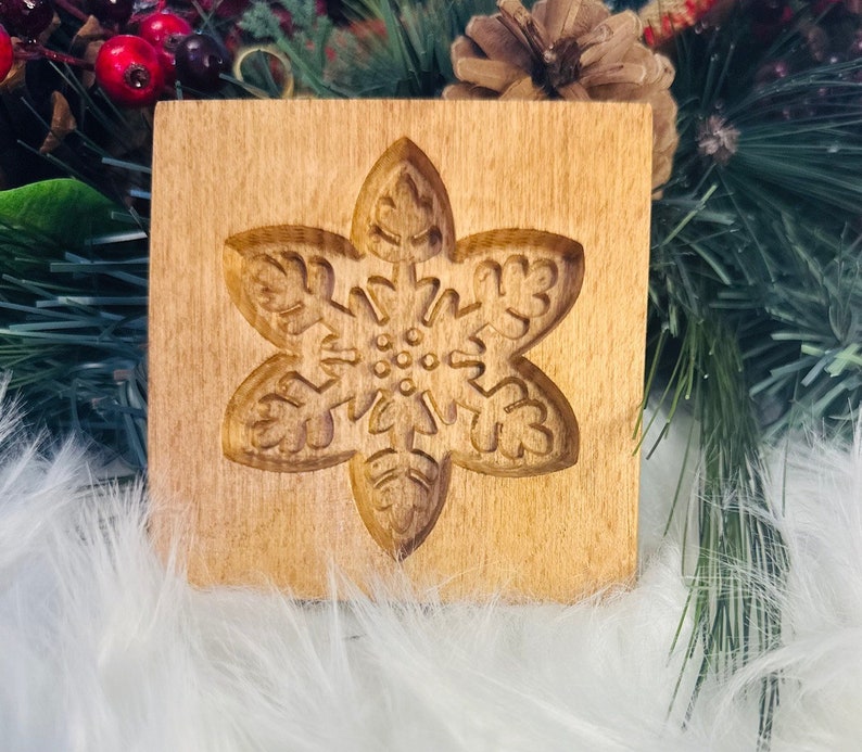 May include: A wooden cookie mold with a carved snowflake design. The mold is square and made of light-colored wood. The snowflake is detailed with intricate patterns, perfect for holiday baking.