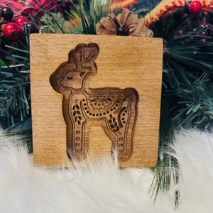 May include: A wooden square with a carved reindeer design. The reindeer is detailed with decorative patterns and is set against a light brown background. The carving is a darker shade of brown, creating a contrast. The item is likely a decorative piece.