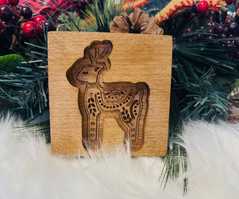 May include: A wooden square with a carved reindeer design. The reindeer is detailed with decorative patterns and is set against a light brown background. The carving is a darker shade of brown, creating a contrast. The item is likely a decorative piece.