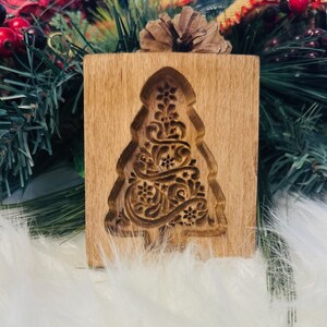 May include: A wooden Christmas tree cookie mold with an intricate floral design carved into the surface. The mold is rectangular with a tree-shaped cutout. The wood has a light brown color.