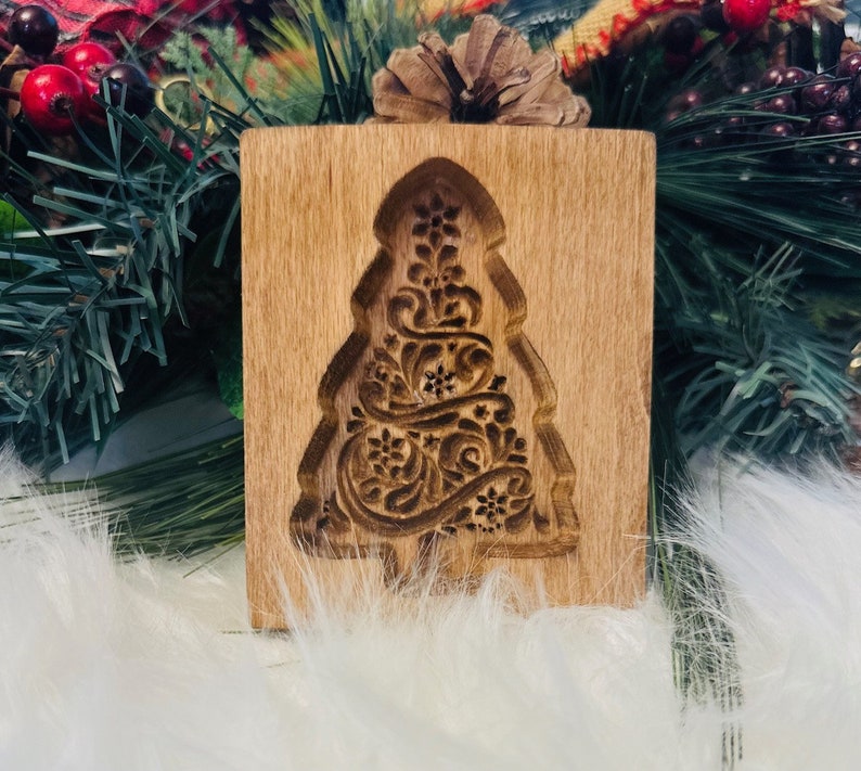 May include: A wooden Christmas tree cookie mold with an intricate floral design carved into the surface. The mold is rectangular with a tree-shaped cutout. The wood has a light brown color.