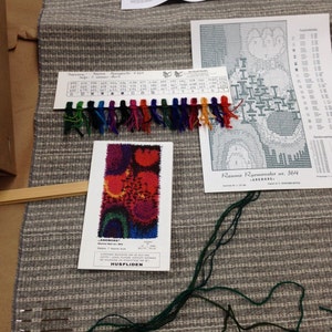 May include: A gray and white striped woven textile with various craft supplies. Includes a color swatch with multiple colored threads, a pattern, and a booklet. The textile is likely for a rug or tapestry project.