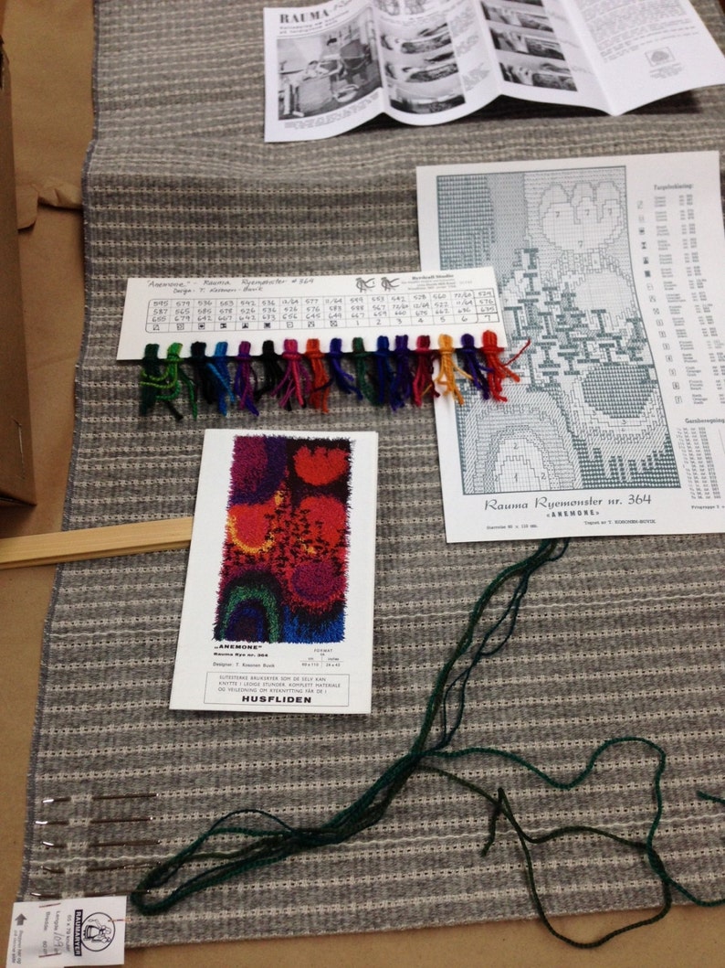 May include: A gray and white striped woven textile with various craft supplies. Includes a color swatch with multiple colored threads, a pattern, and a booklet. The textile is likely for a rug or tapestry project.