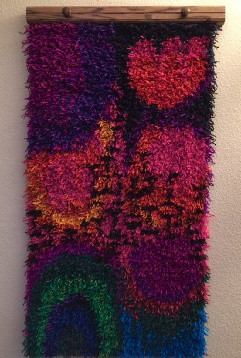 May include: A vibrant, textured wall hanging with a wooden rod. The design features a mix of colors, including purple, pink, orange, and green, arranged in a geometric pattern. The shaggy texture adds depth and visual interest to the artwork.