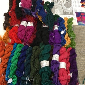 May include: A collection of colorful yarn skeins in various shades, including red, orange, green, blue, and purple. The yarn is arranged on a white surface, with a pattern and color chart visible in the background. A measuring tape is also present.