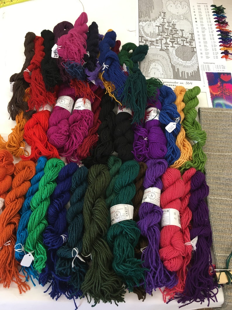 May include: A collection of colorful yarn skeins in various shades, including red, orange, green, blue, and purple. The yarn is arranged on a white surface, with a pattern and color chart visible in the background. A measuring tape is also present.