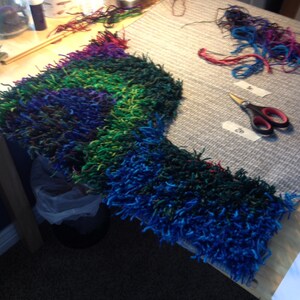 May include: A colorful, handmade textile project in progress, featuring a vibrant mix of blue, green, and purple yarn. The piece is on a wooden table, with scissors and loose yarn nearby. The project appears to be a rug or a similar item.