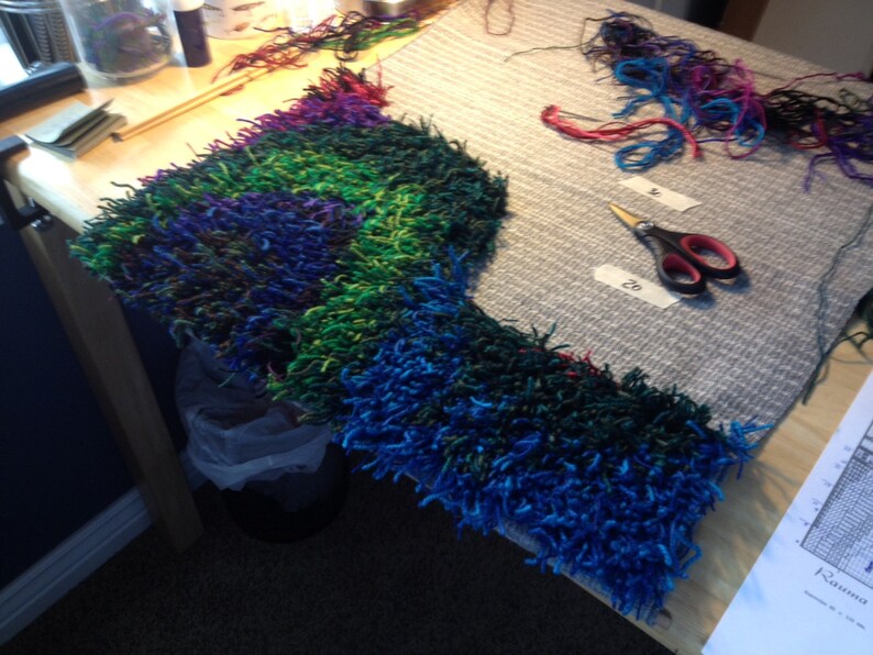 May include: A colorful, handmade textile project in progress, featuring a vibrant mix of blue, green, and purple yarn. The piece is on a wooden table, with scissors and loose yarn nearby. The project appears to be a rug or a similar item.
