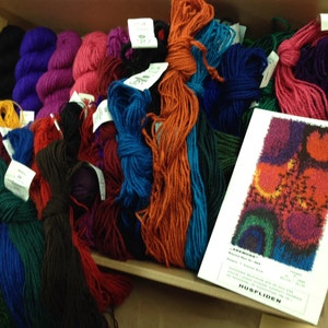 May include: A cardboard box filled with colorful yarn skeins in shades of purple, pink, blue, orange, and green. A white card with a colorful abstract design and text is visible. The yarn is likely for knitting or other fiber crafts.