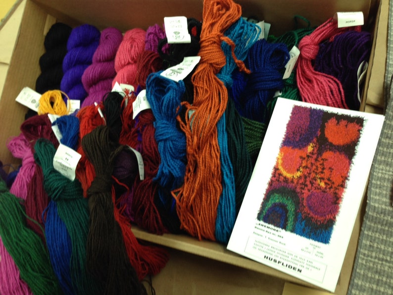 May include: A cardboard box filled with colorful yarn skeins in shades of purple, pink, blue, orange, and green. A white card with a colorful abstract design and text is visible. The yarn is likely for knitting or other fiber crafts.