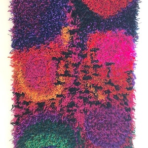 May include: A rectangular, shaggy textile art piece with a vibrant color palette. The artwork features a mix of colors, including red, pink, orange, purple, green, and blue. The word "Anemone" is printed at the bottom.