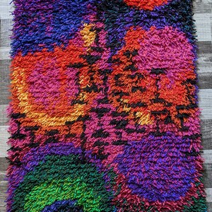 May include: A vibrant, rectangular shag rug with a colorful abstract design. The rug features a mix of colors, including red, pink, purple, green, and blue. The texture is thick and shaggy, creating a cozy and visually appealing piece.
