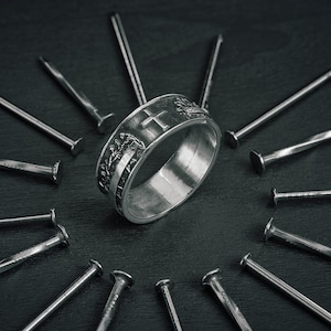 Può includere: Un anello d'argento con un design a croce circondato da un cerchio di chiodi d'argento su una superficie nera.
