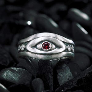 May include: A silver ring featuring a central eye-shaped motif with a small, round, deep red gemstone set in the middle. The band is accented with small, clear, round stones embedded along the sides of the eye design, creating a detailed look.