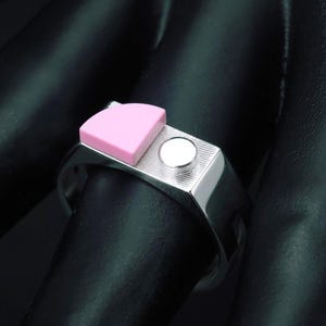 May include: A silver-toned metal ring featuring a flat, rectangular top surface with a textured finish. The ring is adorned with a small, light pink semi-circular accent piece and a tiny, raised silver-toned circular stud positioned next to it.