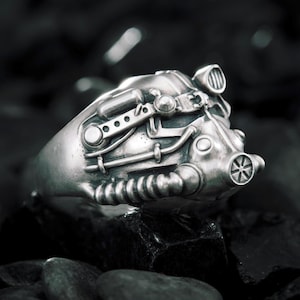 Puede incluir: Un anillo de metal plateado con un diseño esculpido y detallado de un casco futurista que presenta un filtro circular prominente, tubos laterales y un acabado liso y pulido que refleja la luz sobre su superficie compleja y mecánica.