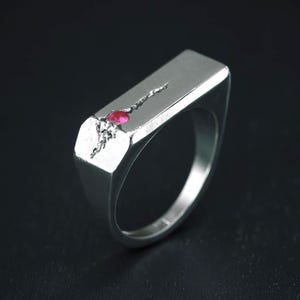 May include: A silver signet ring featuring a flat rectangular top surface. The ring is accented with a small, round, vibrant pink gemstone embedded near the edge, accompanied by a delicate, textured, vine-like silver engraving extending outward.