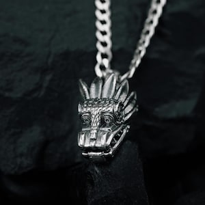 May include: A silver-toned metal pendant shaped like a stylized dragon head with intricate scale detailing and a prominent crest, suspended from a matching silver-toned curb chain necklace. The pendant features a wide snout and defined eyes.