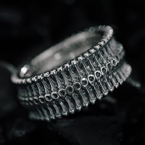 Peut inclure: Une bague en argent avec un design unique composé d'une série de petites formes rondes et allongées. La bague est posée sur un fond sombre et texturé.