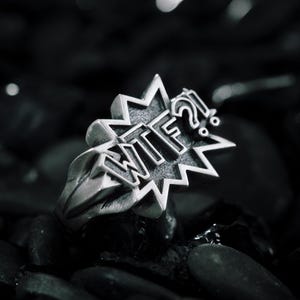 Peut inclure: Une bague en métal argenté présentant un motif de bulle de bande dessinée avec le texte "WTF?!" en relief sur le devant. La bague possède une finition polie avec des zones en creux assombries pour souligner le lettrage et les bords dentelés.