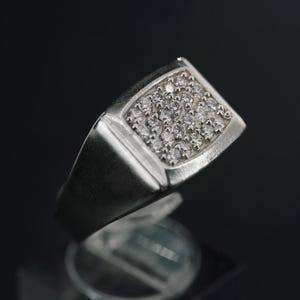 May include: A silver ring with a square face set with numerous small, clear stones. The ring is displayed on a clear, geometric stand against a dark background. The ring's band is thick and solid.