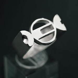 May include: A silver ring with a unique candy-shaped design. The ring features a central circular element with a horizontal bar and two stylized end pieces. The ring is displayed on a clear, square stand against a dark background.