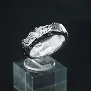 Puede incluir: Un anillo de tono plateado oscuro que presenta una superficie muy texturizada, agrietada e irregular. La banda tiene un aspecto rugoso y desgastado con fisuras profundas en el exterior, mientras que el interior es liso y pulido.