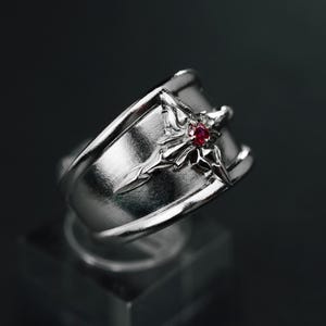 Könnte beinhalten: Ein silberner Ring mit Sternmuster und einem kleinen roten Edelstein in der Mitte. Der Ring wird auf einem transparenten Ständer vor dunklem Hintergrund präsentiert. Das Band des Rings ist breit und hat eine polierte Oberfläche.