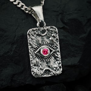 Könnte beinhalten: Silberne Halskette mit Anhänger, rechteckiger Anhänger mit strukturierter Oberfläche. Das Design zeigt ein Auge mit rotem Zentrum, umgeben von Sonnenstrahlen und Monden. Die Kette hat dicke Glieder.