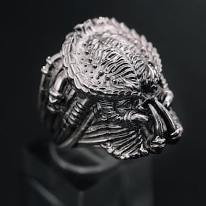 May include: A detailed silver ring featuring the likeness of a Predator's head. The ring showcases intricate details of the creature's face, including mandibles and textured skin. The ring is displayed on a clear stand against a dark background.
