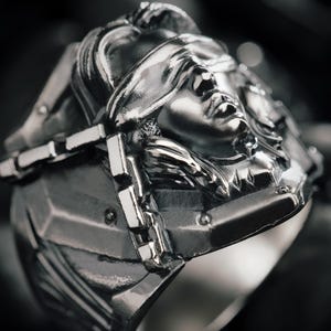 Puede incluir: Anillo plateado detallado con un rostro con los ojos vendados, posiblemente la Justicia. El anillo tiene un diseño geométrico en la banda y un elemento en forma de cadena en la cara. La estética general es oscura y gótica.
