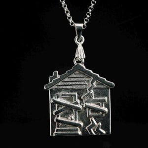 Puede incluir: Un collar de plata con un colgante en forma de casa destartalada. La casa tiene ventanas tapiadas y un techo agrietado.