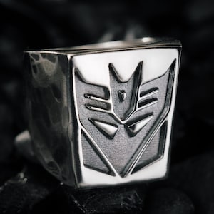 Puede incluir: Un anillo plateado con una cara cuadrada que presenta el símbolo de los Decepticons. El anillo tiene una textura martillada y un acabado pulido, mostrando el emblemático emblema de la franquicia Transformers.