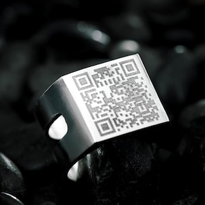 Puede incluir: Un anillo de metal plateado con una superficie superior plana y cuadrada grabada con un patrón de código QR negro detallado. La banda tiene una construcción ancha y sólida con un acabado liso y pulido y un diseño de corte lateral.