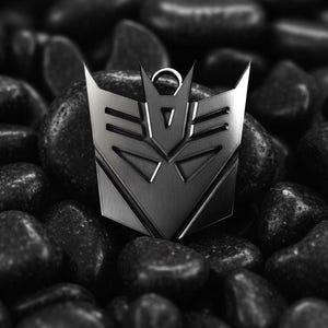 May include: A silver pendant displaying the Decepticon symbol, a geometric emblem with a central triangle and sharp angles. The pendant is set against a backdrop of dark, smooth stones, creating a striking contrast.