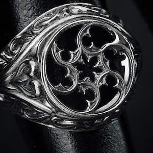 May include: Close-up of a silver ring with an ornate, gothic design. The ring has a central openwork pattern surrounded by detailed scrollwork and floral motifs. The ring is set against a dark background.