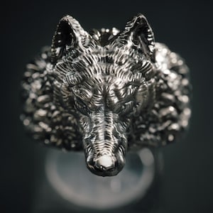 May include: A silver ring with a detailed wolf's head design. The ring displays textured details, capturing the wolf's fur and facial features. The ring's metallic surface is highlighted against a dark backdrop, showcasing its craftsmanship.