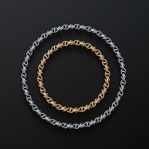May include: Two chain necklaces, one silver and one gold, arranged in concentric circles against a black background. The chains have a repeating link design. The gold chain is smaller and inside the silver chain.