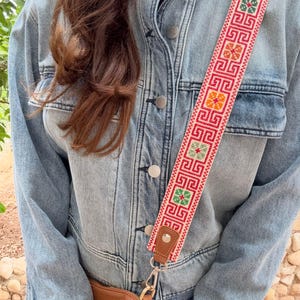 May include: A denim jacket with a decorative, patterned strap in red, orange, green, and white. The strap is attached to a brown leather bag with a gold-colored clasp. The jacket has silver buttons.