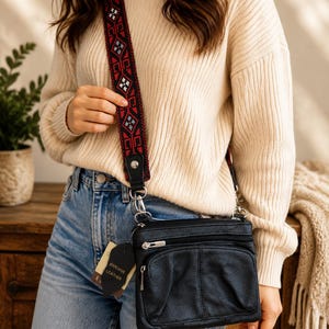 Puede incluir: Un bolso bandolera de cuero negro con superficie texturizada, múltiples compartimentos con cremallera y una correa ancha y extraíble con un patrón geométrico rojo y negro. Una pequeña etiqueta negra con la inscripción Genuine Leather está sujeta al lateral.