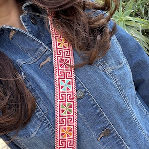 May include: A denim jacket with a decorative strap. The strap is red with a geometric pattern in white, blue, orange, and green. The jacket is a medium wash denim with silver buttons.