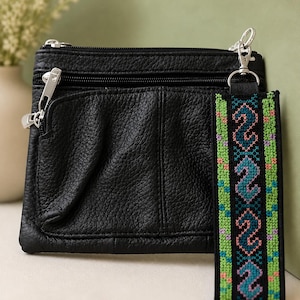 May include: Black leather crossbody bag with multiple zippered compartments and a decorative embroidered keychain. The keychain features a geometric pattern in shades of green, blue, and pink. The bag has silver-toned hardware.