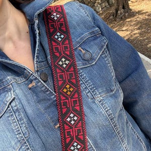 May include: A denim jacket with a decorative strap. The strap is black with a repeating geometric pattern in red, white, and yellow. The jacket is a medium wash blue with button closures and pockets.