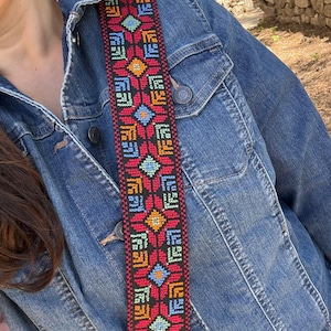 Puede incluir: Una colorida correa tejida con un patrón geométrico en rojo, negro, azul, amarillo y naranja. La correa se lleva sobre una chaqueta vaquera. El diseño de la correa presenta formas florales y de diamante repetidas.
