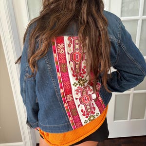 May include: A denim jacket with a colorful embroidered panel on the back. The panel features a white background with red, pink, and green floral and geometric designs. The jacket has a classic collar and button-up front.
