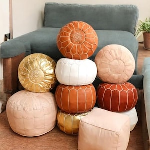 May include: A collection of decorative leather poufs in various colors, including tan, white, gold, and rust. The poufs are round and square, with stitched geometric patterns. They are stacked in a visually appealing arrangement.