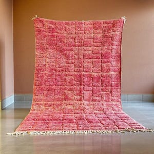 Peut inclure: Un grand tapis rectangulaire rose avec un motif géométrique texturé. Le tapis a un poil épais et une frange le long du bord inférieur. Le tapis est présenté sur un fond neutre.