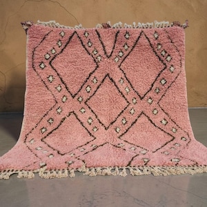 May include: A pink Moroccan rug featuring a geometric diamond design in dark brown. The rug has a soft, textured appearance and is trimmed with fringe along the edges. The rug is displayed on a grey surface.