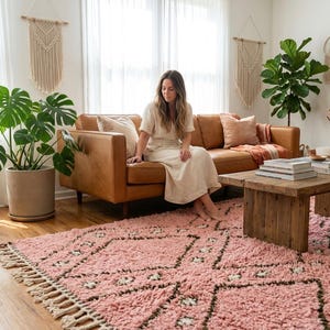 Op de afbeelding: Een pluche, lichtroze vloerkleed met een donkerbruin geometrisch ruitpatroon en decoratieve franjes langs de randen. Het kleed heeft een dikke, hoogpolige textuur en ligt op een houten vloer in een woonkamer.